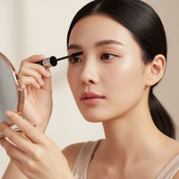 A woman applying My Glam Room BOB Super Curl Mascara to her eyelashes while looking into a small handheld mirror.